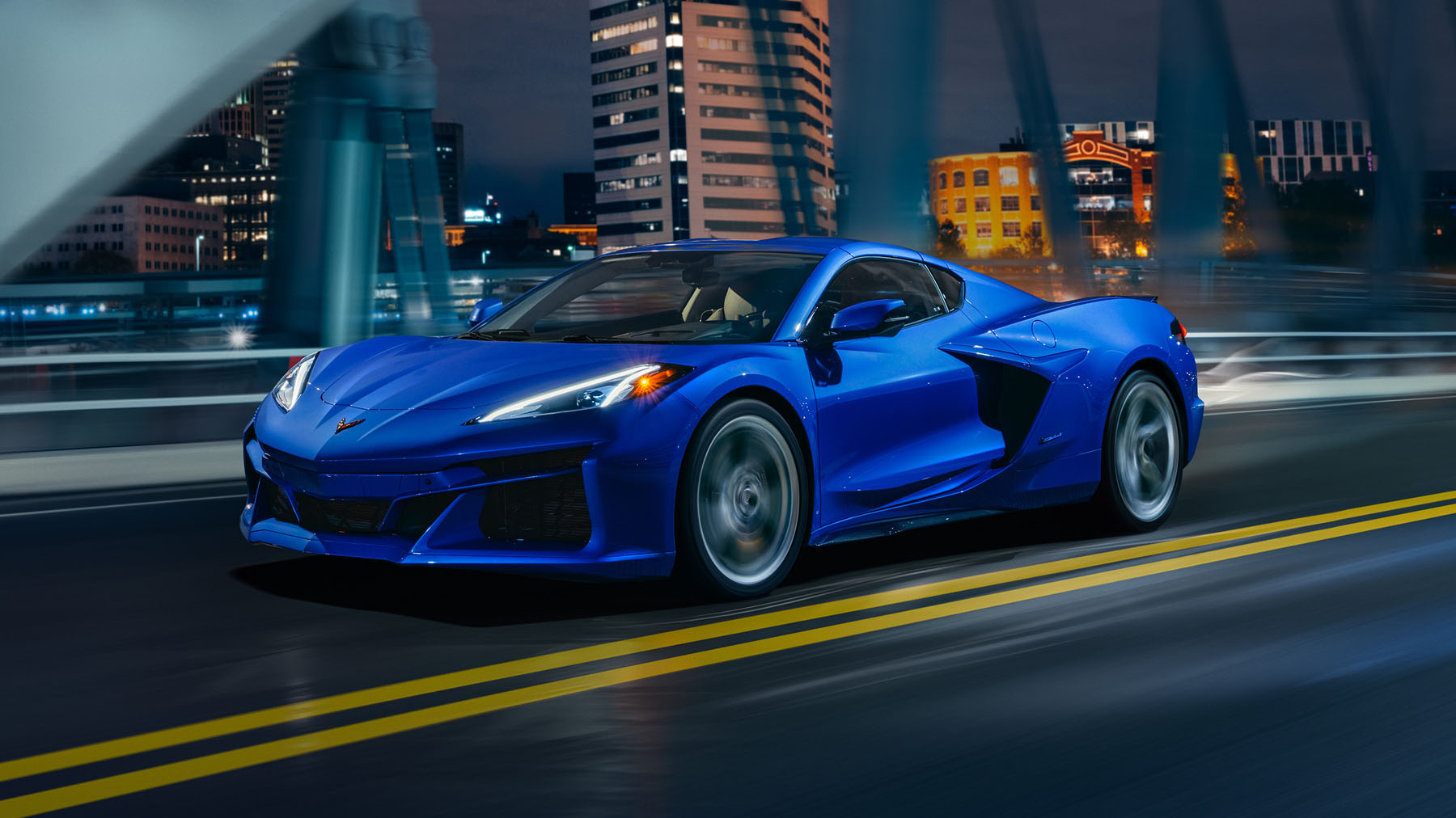 Front 3/4 view of 2024 Chevrolet Corvette E-Ray 3LZ coupe in Riptide Blue, driving over a bridge in front of a city. Pre-production model shown. Actual production model may vary. Model year 2024 Corvette E-Ray available 2023.